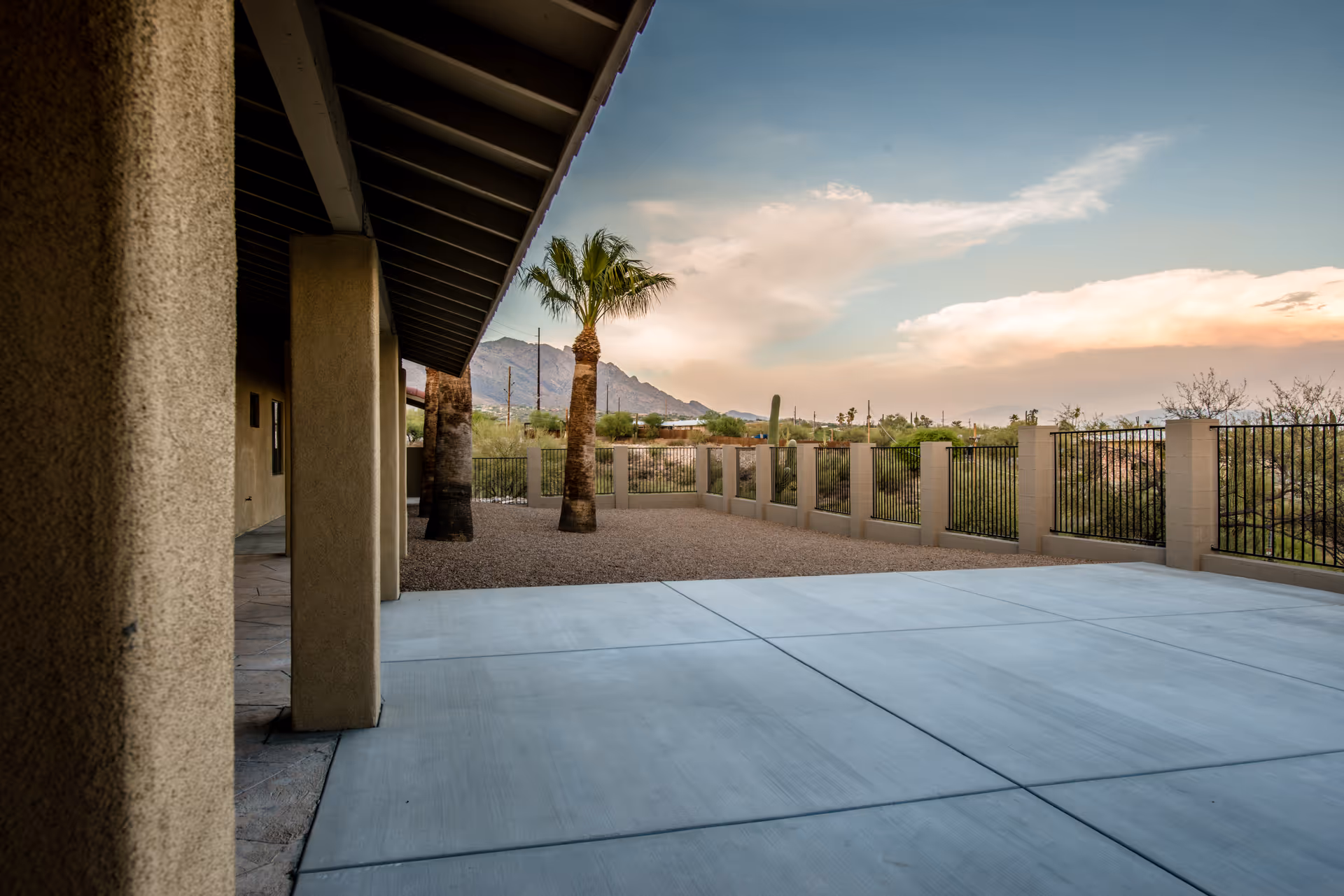 Outdoor patio area with concrete flooring and a covered walkway supported by columns. There are palm trees and desert landscaping with gravel, enclosed by a low wall and metal fencing. Mountains and a partly cloudy sky are visible in the background.