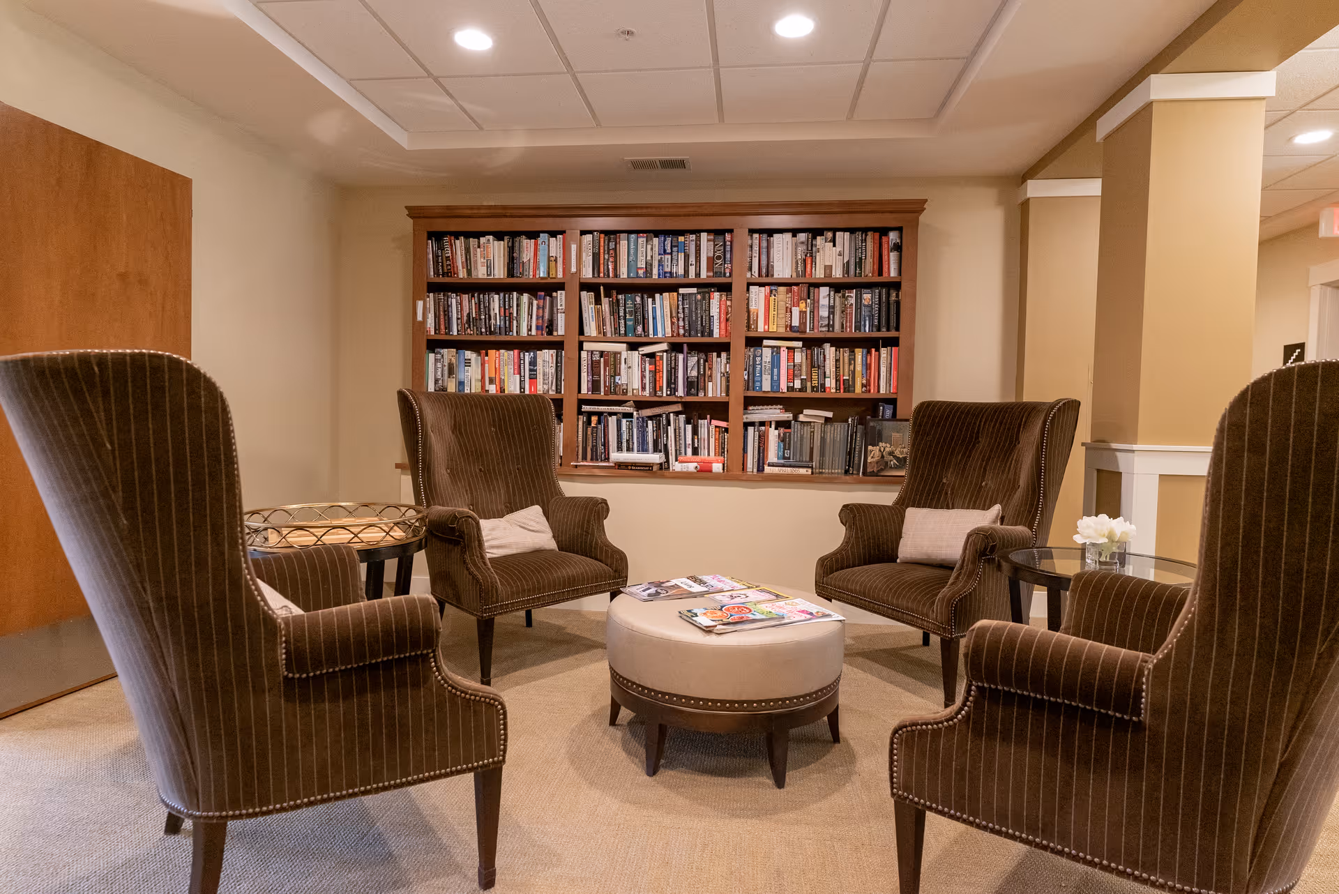 A cozy reading area with four brown upholstered armchairs arranged around a round beige ottoman with magazines on top. Behind the chairs is a built-in wooden bookshelf filled with books. The room has beige walls, carpeted floor, and recessed ceiling lights.