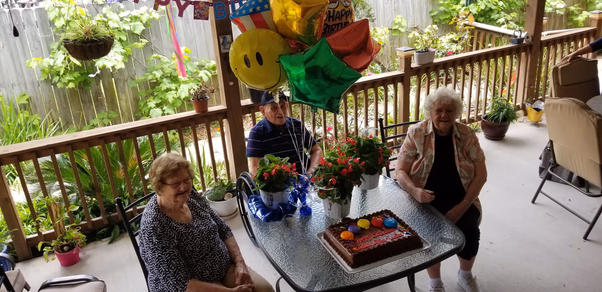 Three elderly people sitting around a glass outdoor table on a covered patio. The table has a chocolate birthday cake with colorful decorations, two potted flowering plants, and a bunch of colorful balloons including a smiley face and stars. The patio is surrounded by a wooden railing and greenery is visible in the background.