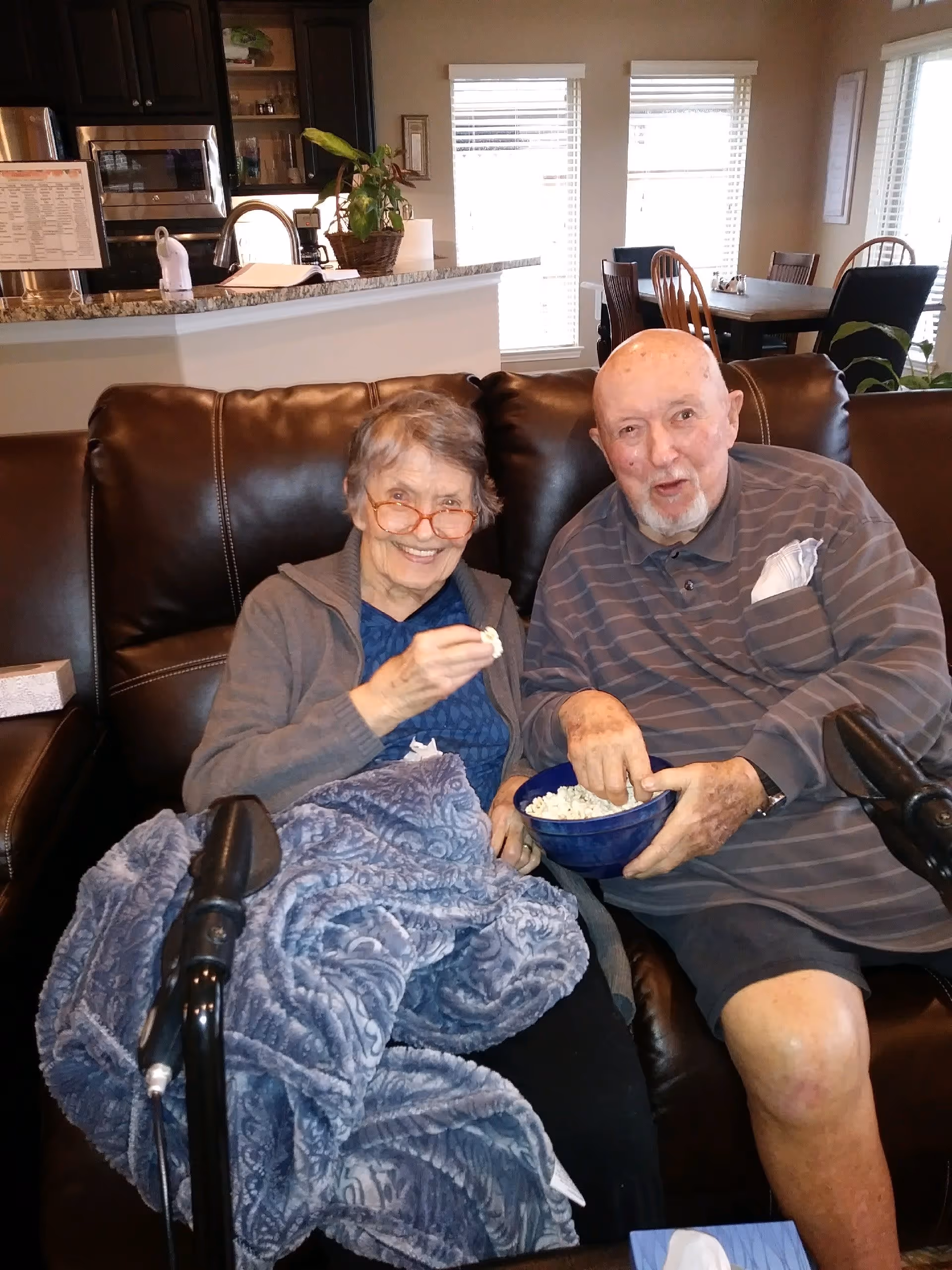 An elderly woman and man sitting together on a dark brown leather couch in a living room. The woman is smiling, wearing glasses and a gray cardigan, with a blue patterned blanket on her lap. The man is wearing a striped polo shirt and shorts, holding a blue bowl of popcorn. Behind them is a kitchen area with a granite countertop and dining table with chairs near windows with blinds.