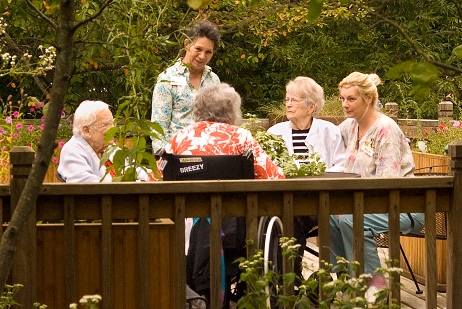 A group of elderly women and two caregivers sitting and standing around a wooden table in a garden area with greenery and flowers, engaging in conversation.