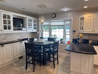 A bright kitchen with white cabinetry and black countertops. There is a round dining table with four black chairs in the center of the room. A large window with a view outside is behind the table, and a clock is mounted above the window. The kitchen features a built-in microwave, a sink, and various kitchen items on the countertops.
