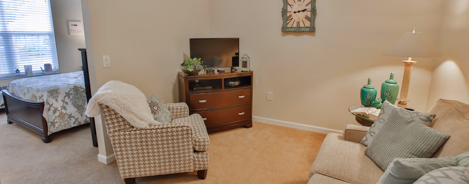Cozy living area with a sofa, armchair, TV on a dresser, and a bed visible in an adjacent bedroom.