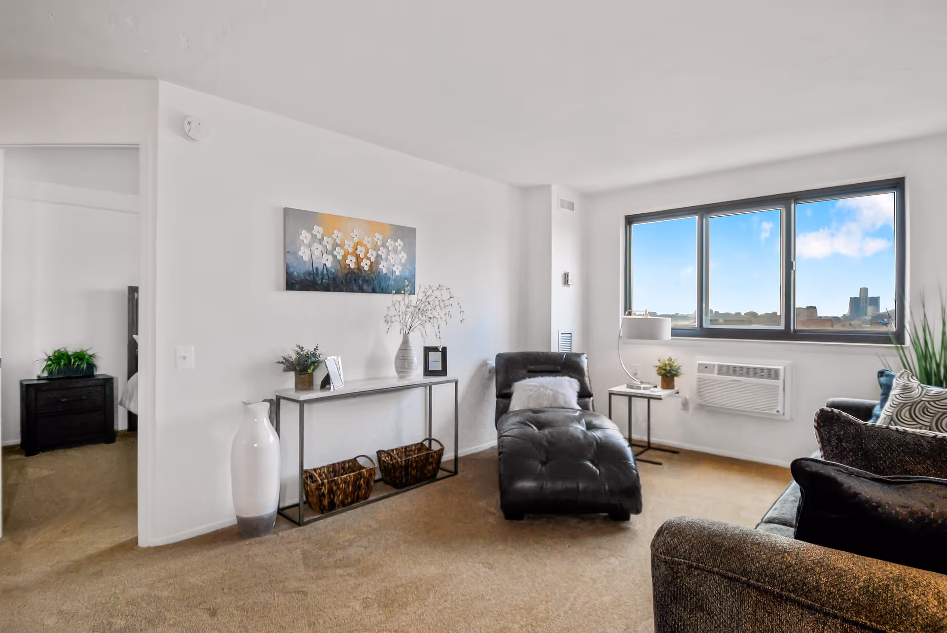 Bright living room with a chaise lounge, sofa, console table, and a large window showing a city view.
