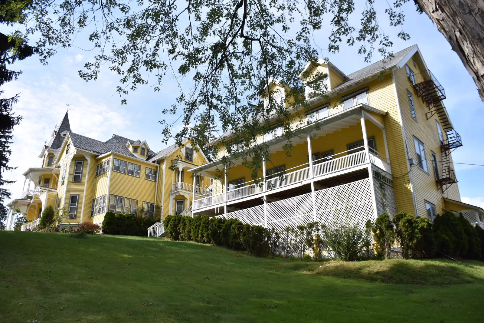 Large yellow Victorian-style senior living building with a wraparound porch on a grassy hill framed by trees.