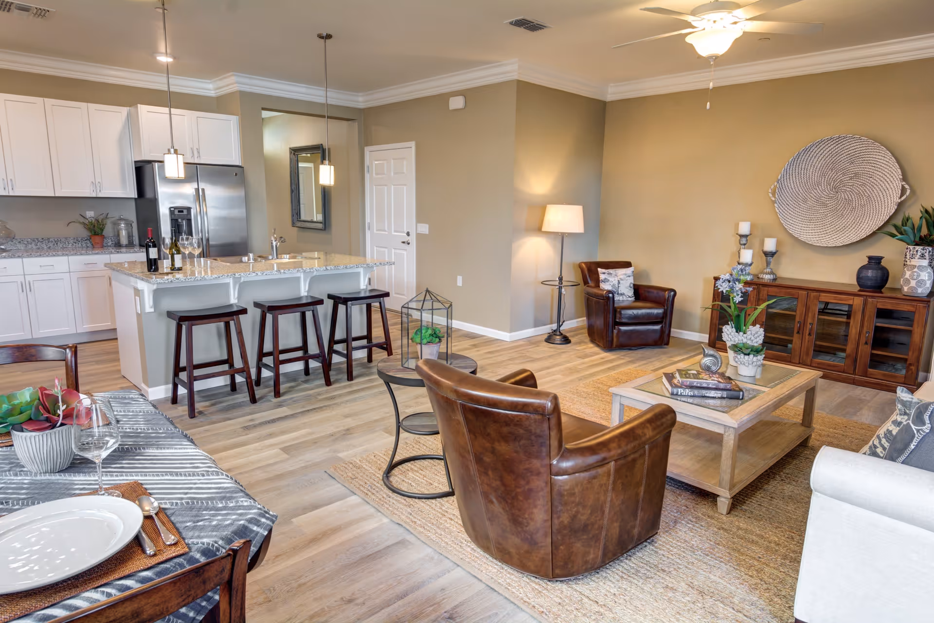 Open concept living area in a senior living facility with a kitchen, dining, and living room. The kitchen features white cabinets, a granite countertop island with three dark wooden stools, and stainless steel appliances. The living room has two brown leather armchairs, a beige sofa, a wooden coffee table with books and plants, a wooden sideboard with decorative items, and a large woven wall decoration. The dining area has a table set with plates, glasses, and cutlery. The room has light wood flooring, beige walls, and ceiling fans with lights.