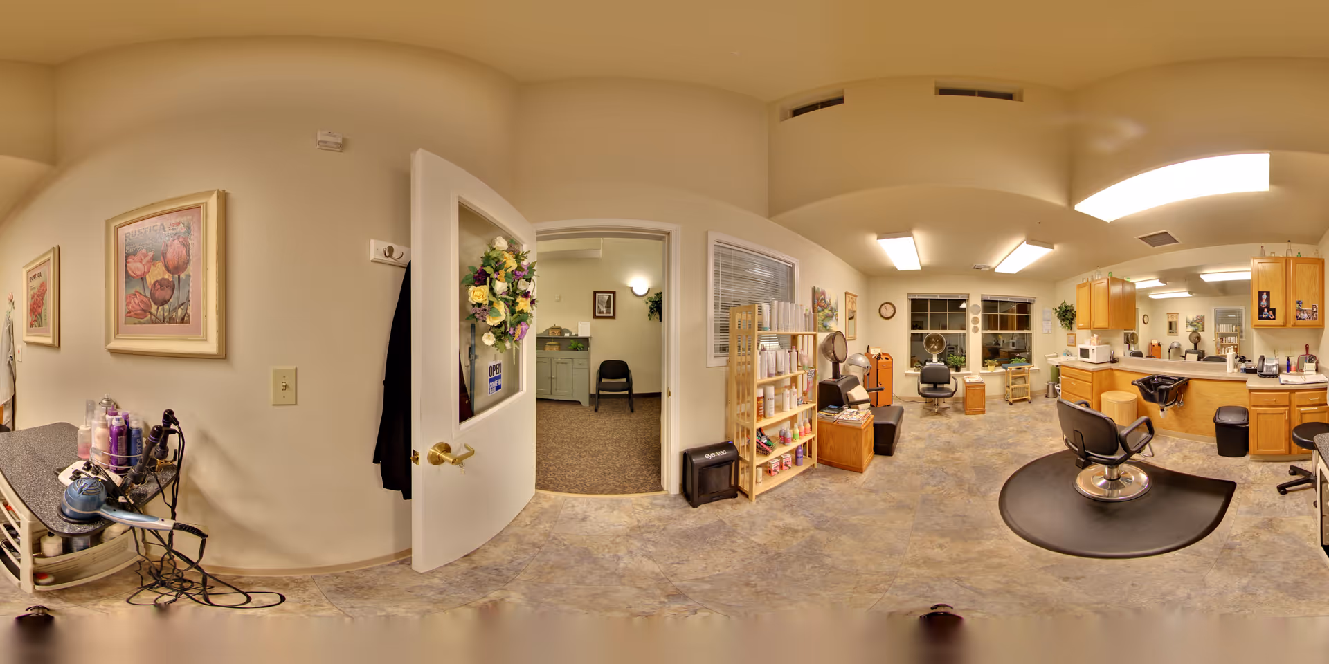 Interior view of a senior living facility's hair salon area with multiple styling chairs, mirrors, and hair care products on shelves. The room has beige walls, tiled flooring, and bright overhead lighting. To the left, there is an open door decorated with a floral wreath leading to another room with a chair and framed pictures on the wall. Various hair styling tools and products are visible on a counter and a small cart.