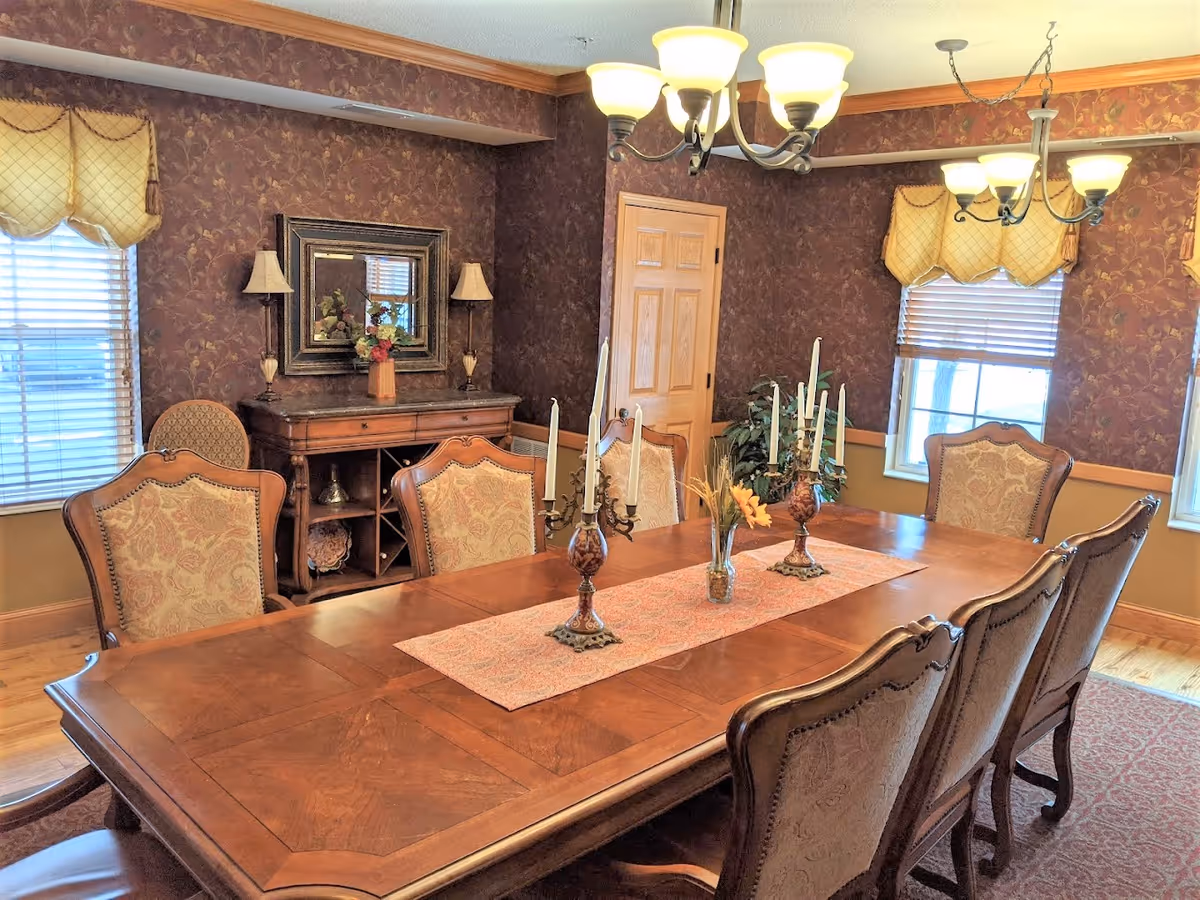 A traditional dining room with a large wooden dining table surrounded by eight upholstered chairs. The table is decorated with two ornate candelabras and a small vase with flowers on a patterned table runner. The room has patterned wallpaper, two windows with yellow valances, a wooden sideboard with a mirror and lamps, and two chandeliers hanging from the ceiling.