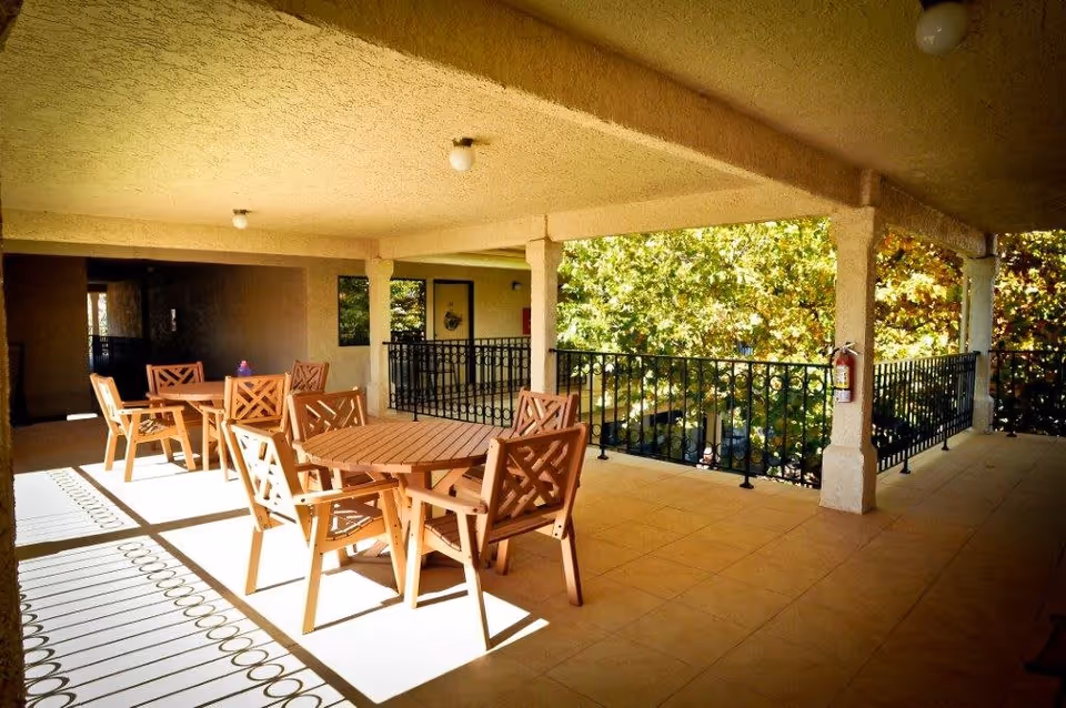 Covered outdoor patio area with multiple wooden tables and chairs arranged for seating. The patio has tiled flooring, beige textured ceiling and walls, black metal railings, and a fire extinguisher mounted on one of the columns. Trees with green foliage are visible beyond the railing.