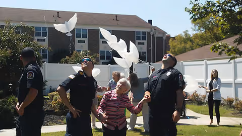 Several people, including firefighters and an elderly woman, are outside in a garden area releasing white dove-shaped balloons into the air near a white fence and residential buildings.