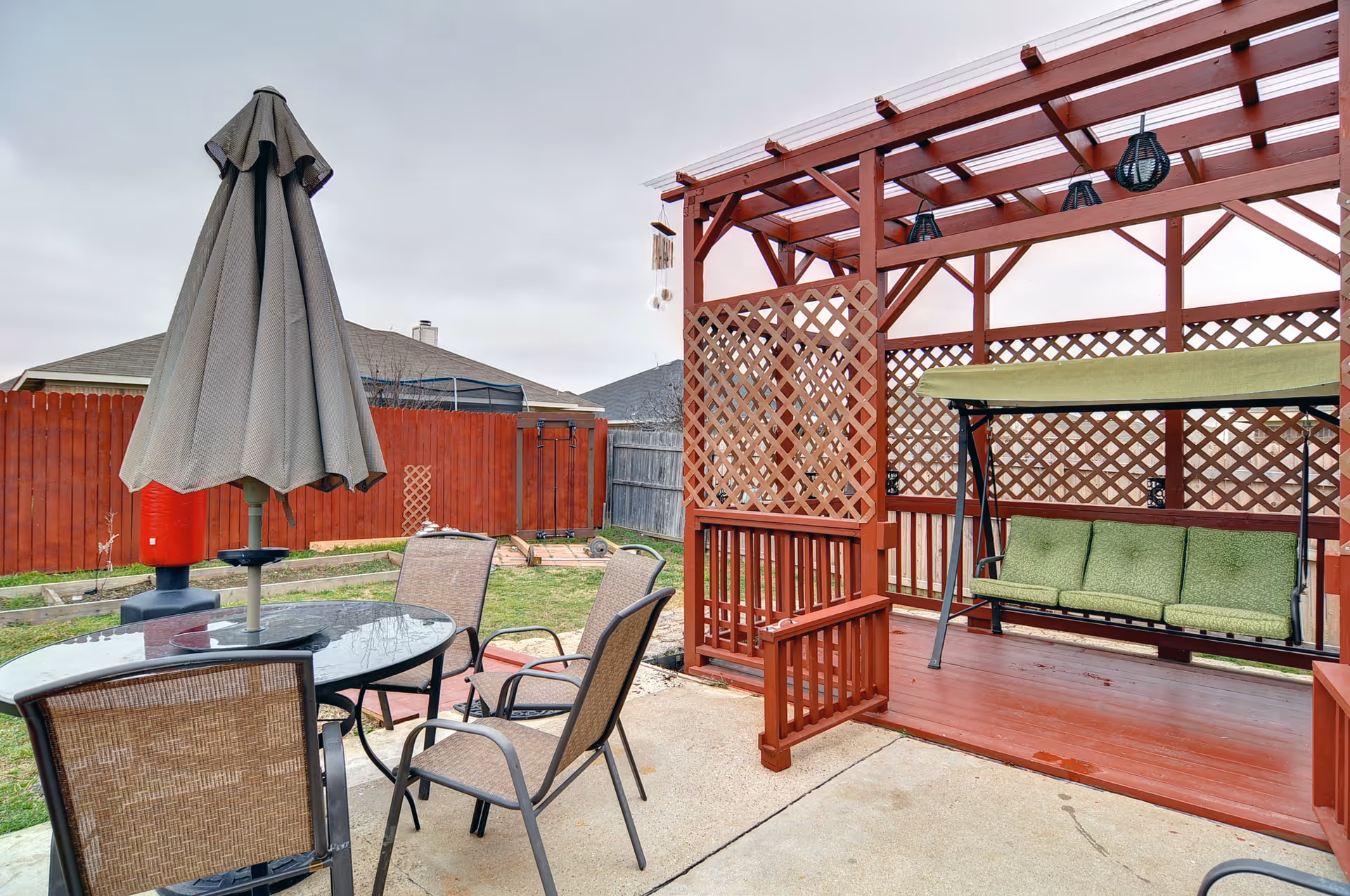 Backyard patio with a red wooden pergola and green-cushioned swing, a round glass table with umbrella and several chairs.