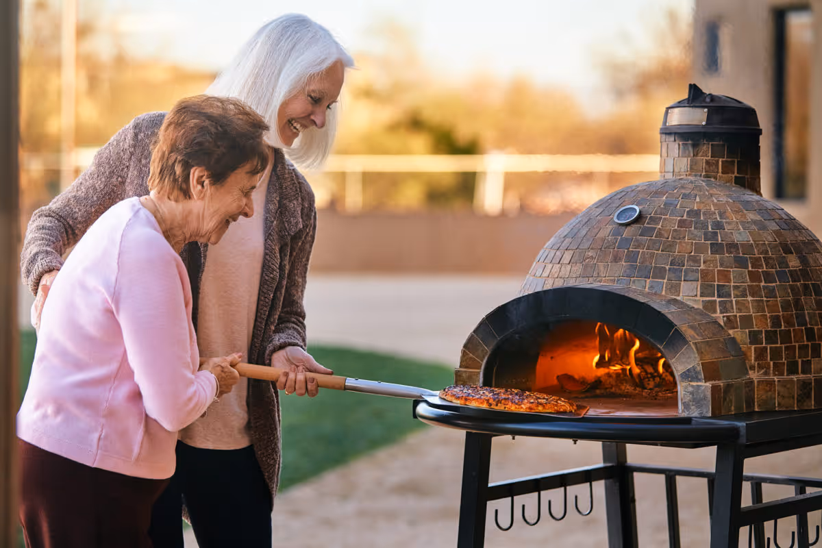 Two elderly women smiling and working together to take a pizza out of a wood-fired outdoor oven using a pizza peel, with a blurred outdoor background.