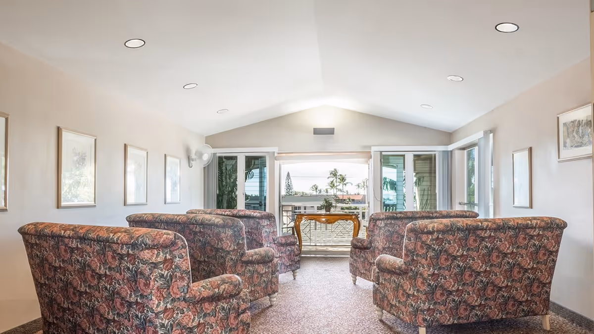 A bright living room with six floral-patterned armchairs arranged in two rows facing a small wooden table near large glass doors that open to a balcony with a view of palm trees and a building outside. The room has beige walls adorned with framed pictures and recessed ceiling lights.