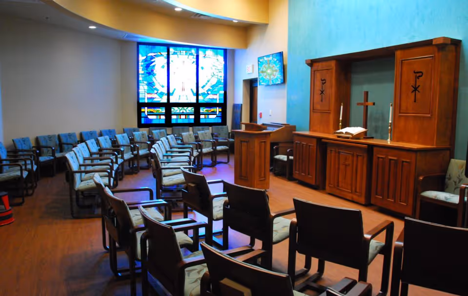 A small chapel-style interior with rows of chairs facing a wooden altar and a stained glass window.