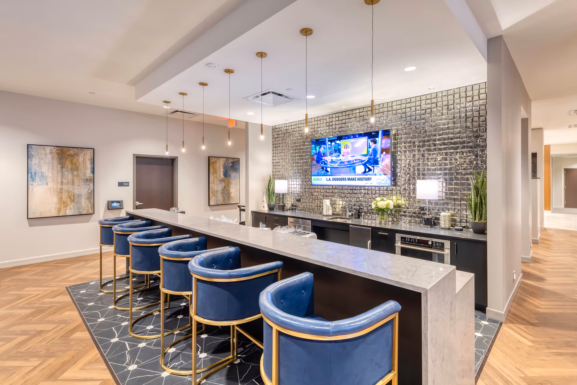 Modern lounge area with a long marble countertop bar and six blue upholstered bar stools with gold frames. Behind the bar is a wall with reflective square tiles, two table lamps, plants, and a mounted flat-screen TV displaying a news segment. The floor has a patterned black rug under the bar stools and wood flooring in the surrounding area. Two abstract paintings hang on the wall near a dark door.