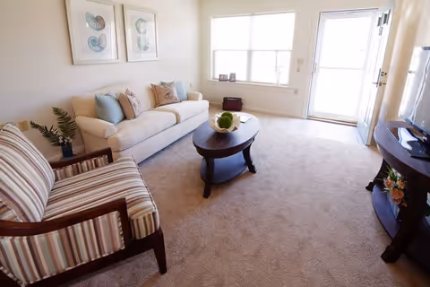 A bright living room with a beige carpet, a white sofa with blue and beige cushions, a striped armchair, a dark wooden oval coffee table with a decorative centerpiece, two framed artworks on the wall, a window letting in natural light, and a door leading outside.
