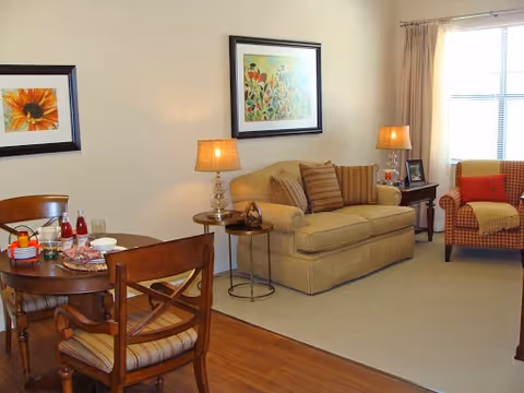 A cozy living room and dining area with a beige sofa, striped cushions, a patterned armchair with a red pillow, two table lamps, framed floral artwork on the walls, a wooden dining table set with plates and drinks, and a window with beige curtains letting in natural light.