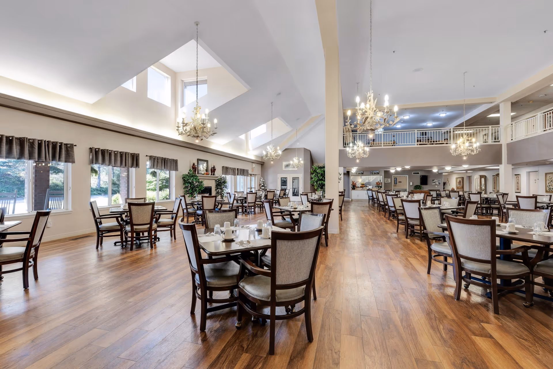 Spacious dining room with wooden floors, multiple tables and chairs set with white cups and plates, large windows with curtains allowing natural light, chandeliers hanging from a high ceiling, and a second-floor balcony overlooking the area.