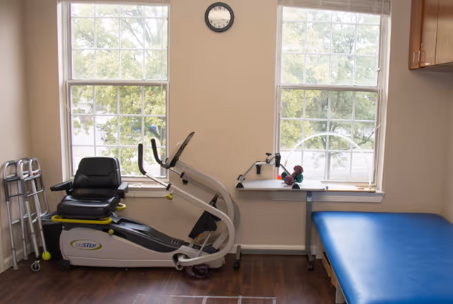 A small exercise room with a recumbent exercise bike, a walker, a blue padded examination or therapy table, a small table with hand weights, and two large windows letting in natural light.