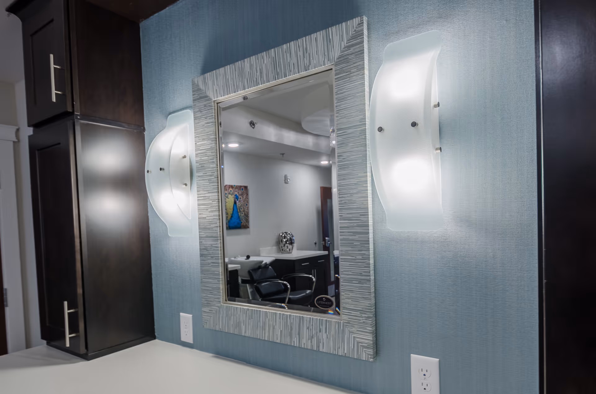 A wall-mounted mirror with a textured silver frame is centered between two modern frosted glass wall sconces. The wall behind is covered in a light blue textured wallpaper. On either side of the mirror are dark wood cabinets with silver handles. The mirror reflects a room with a black chair, a countertop, a decorative vase, and a colorful peacock painting on the wall.