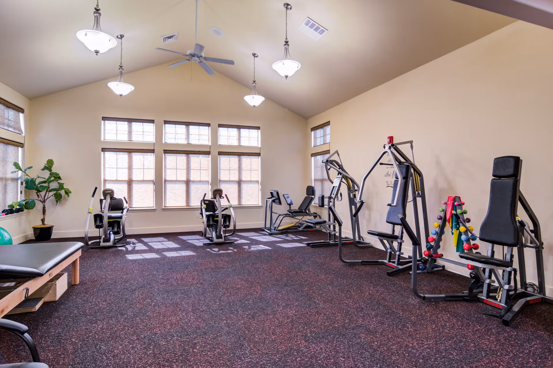 A bright fitness room with exercise equipment including stationary bikes, weight machines, and a rack of colorful dumbbells. The room has large windows with blinds, beige walls, a high ceiling with ceiling fans and hanging lights, and a carpeted floor.