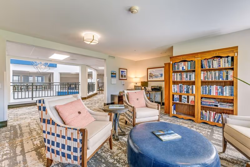 A cozy senior living common area with three cushioned armchairs featuring pink pillows arranged around a round blue ottoman with a book on top. There is a small round side table between two chairs. In the background, a wooden bookshelf filled with books is against the wall, and a desk with a lamp and computer is visible. The space has carpeted flooring and overlooks an open hallway with railings and large windows.