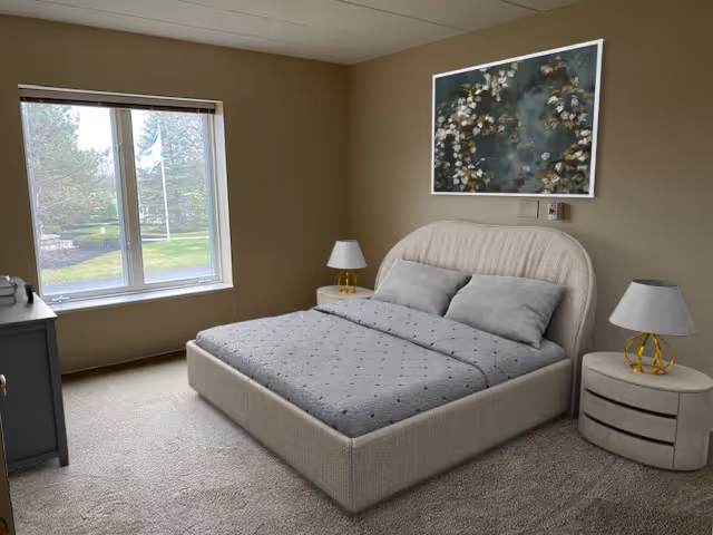 A neatly made bed with a light gray upholstered headboard and matching bedding in a beige carpeted room. There are two bedside tables, each with a lamp featuring a white shade and gold base. A large window on the left side lets in natural light and shows a green outdoor area. A framed floral artwork hangs on the wall above the bed.