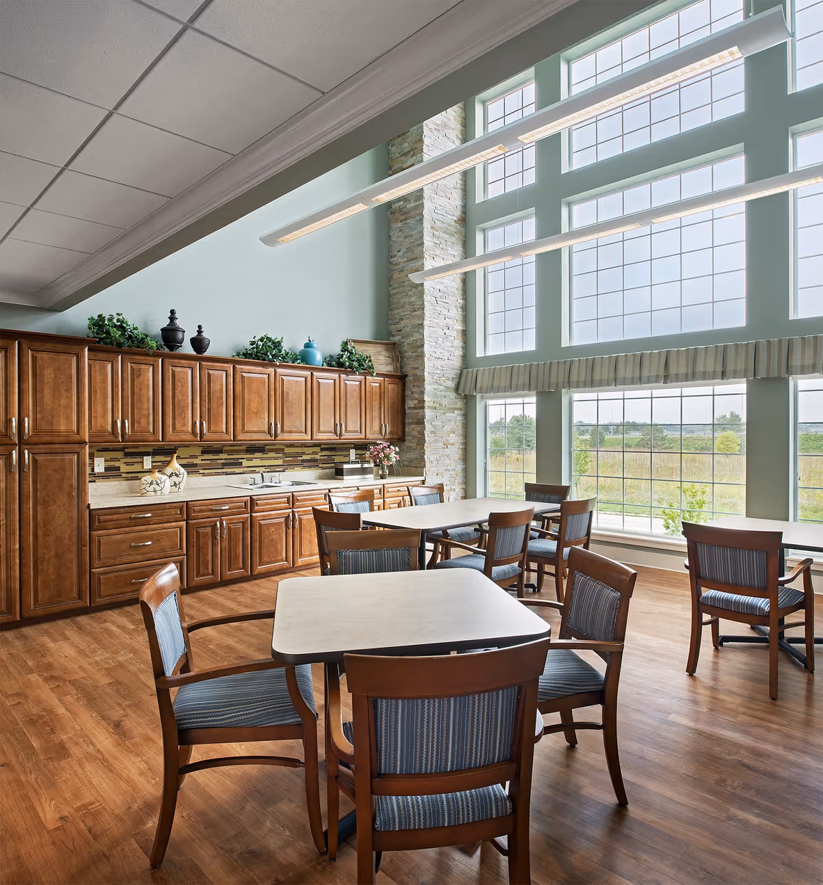 A bright dining area with large floor-to-ceiling windows letting in natural light. The room features wooden tables and chairs with blue cushioned seats, a long countertop with wooden cabinets above and below, decorative plants and vases on top of the cabinets, and a stone accent wall between the windows.