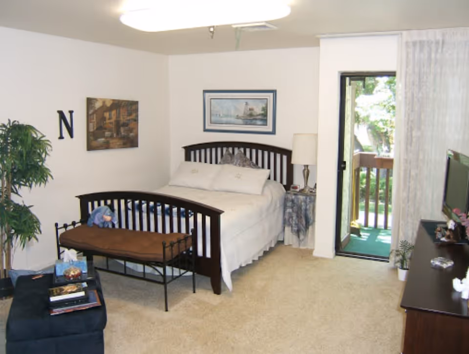 Bright bedroom with a bed and bench, dresser and TV, a potted plant, and a sliding door leading to a small balcony.