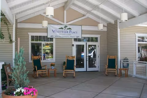 Covered outdoor seating area with four wooden rocking chairs and small tables, potted plants with flowers, and a sign above the entrance doors that reads 'Whittier Place Welcomes You'.