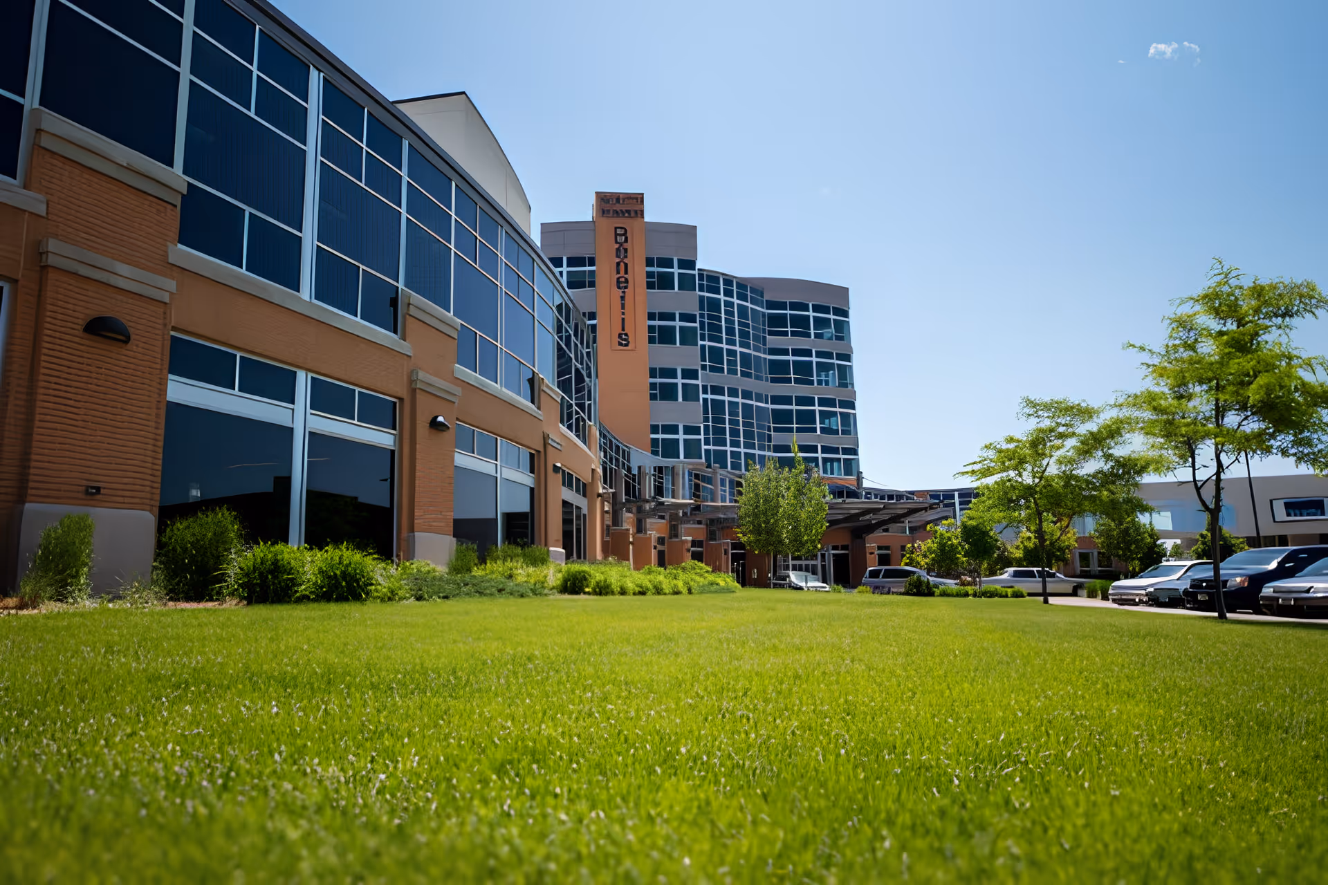 Exterior view of a modern multi-story building with large windows, a well-maintained green lawn in the foreground, and several trees and parked cars on the right side under a clear blue sky.