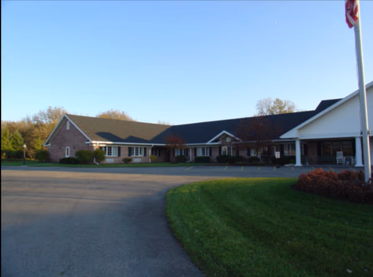Exterior view of a single-story senior living facility building with a brick facade and a dark shingled roof, surrounded by a paved driveway and green lawn under a clear blue sky.