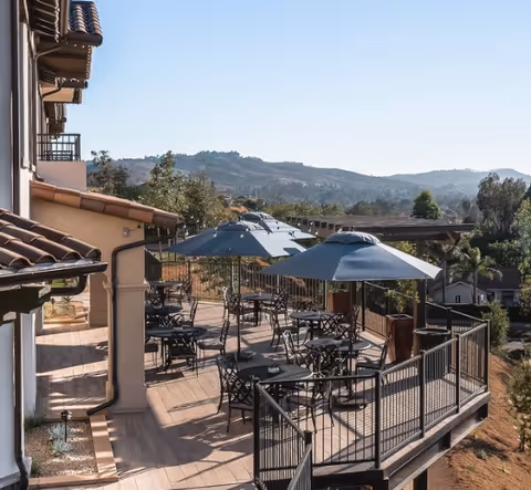 Outdoor patio area at a senior living facility with multiple black metal tables and chairs, shaded by large blue umbrellas. The patio overlooks a scenic view of hills and trees under a clear blue sky.