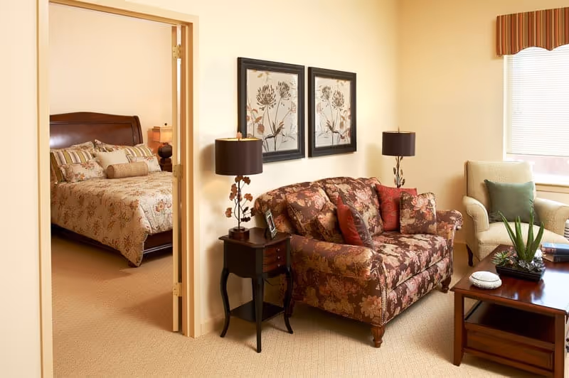 Living area with a floral-patterned loveseat, side tables and lamps, a coffee table, and a bedroom visible through an open doorway.