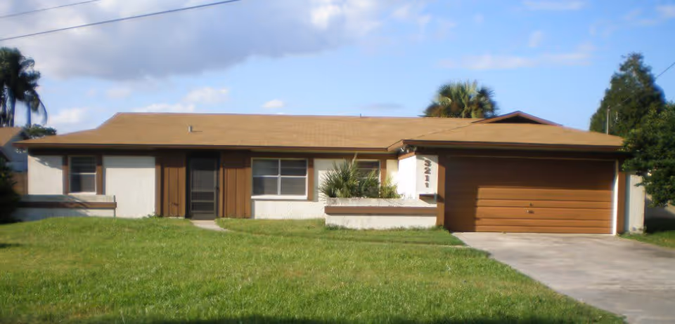 Single-story ranch-style house with a front lawn, attached two-car garage, and palm trees in the background.
