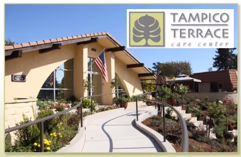 Exterior view of Tampico Terrace Care Center showing a beige building with a tiled roof, a curved walkway with handrails leading to the entrance, an American flag, and landscaped garden beds with flowers and shrubs.