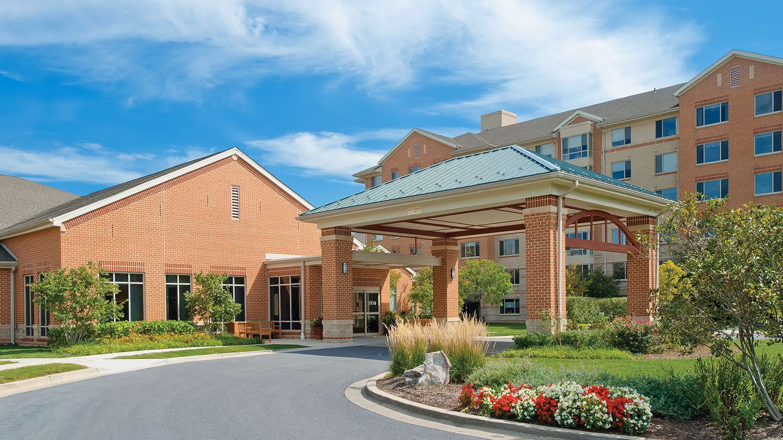 Exterior view of Riderwood Senior Living Community showing a brick building with a covered entrance, landscaped garden with flowers and shrubs, and a clear blue sky.