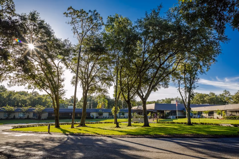 Exterior view of Solaris HealthCare Plant City facility with a single-story building surrounded by green lawns and tall trees under a clear blue sky with the sun shining through the branches.