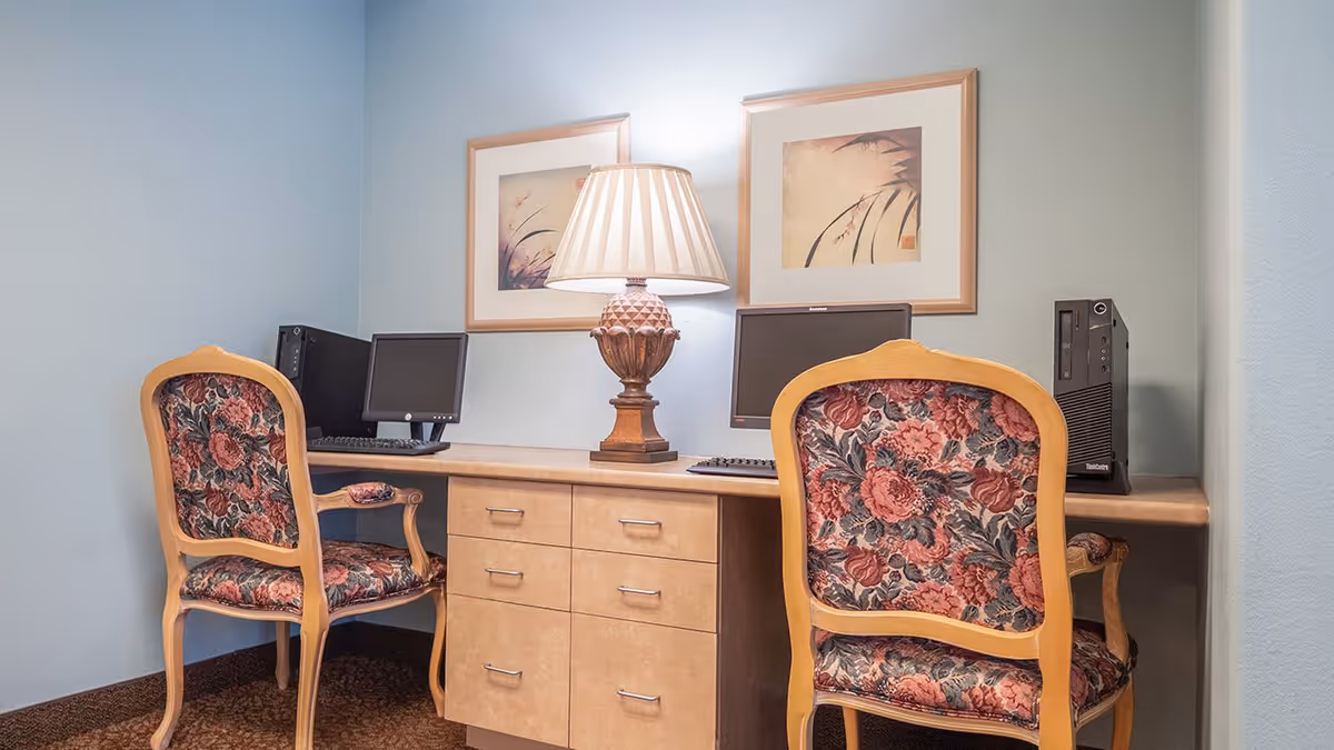 A small office or computer area with two vintage-style wooden chairs upholstered in floral fabric, a wooden desk with drawers, two desktop computers, and a decorative table lamp in the center. Two framed floral artworks hang on the light blue wall behind the desk.