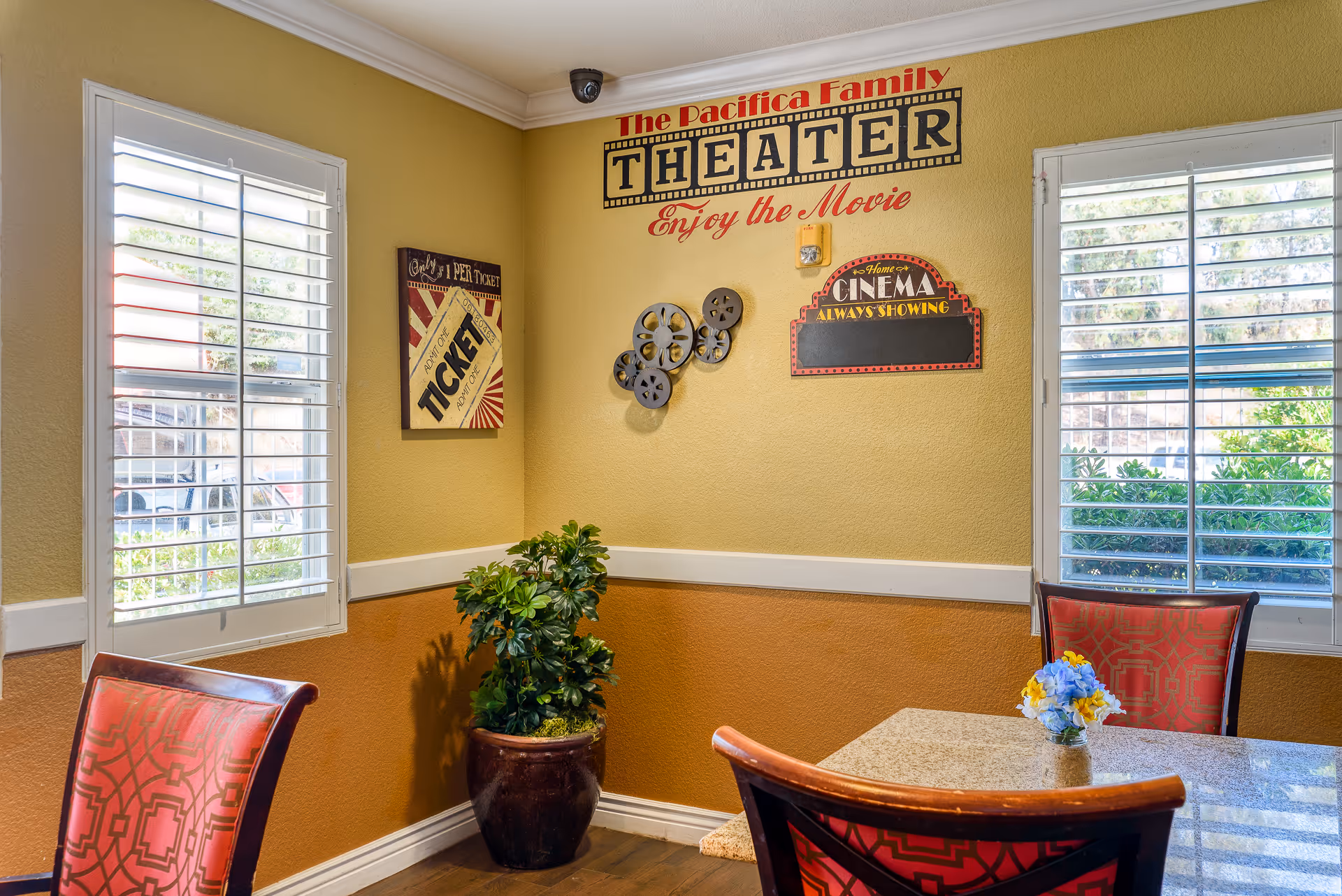 Bright corner dining area with a small table and chairs, a potted plant, shuttered windows and movie-themed wall decor.