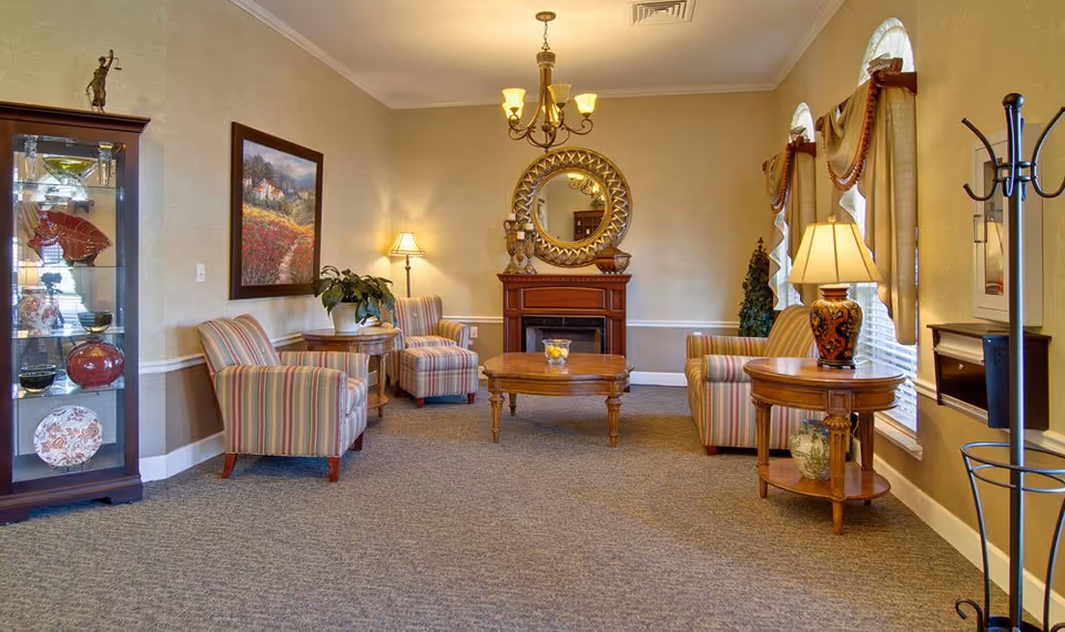 A cozy senior living common area with striped armchairs, wooden side tables with lamps, a wooden coffee table, a decorative fireplace with a round mirror above it, a glass display cabinet with decorative items, and windows with curtains letting in natural light.