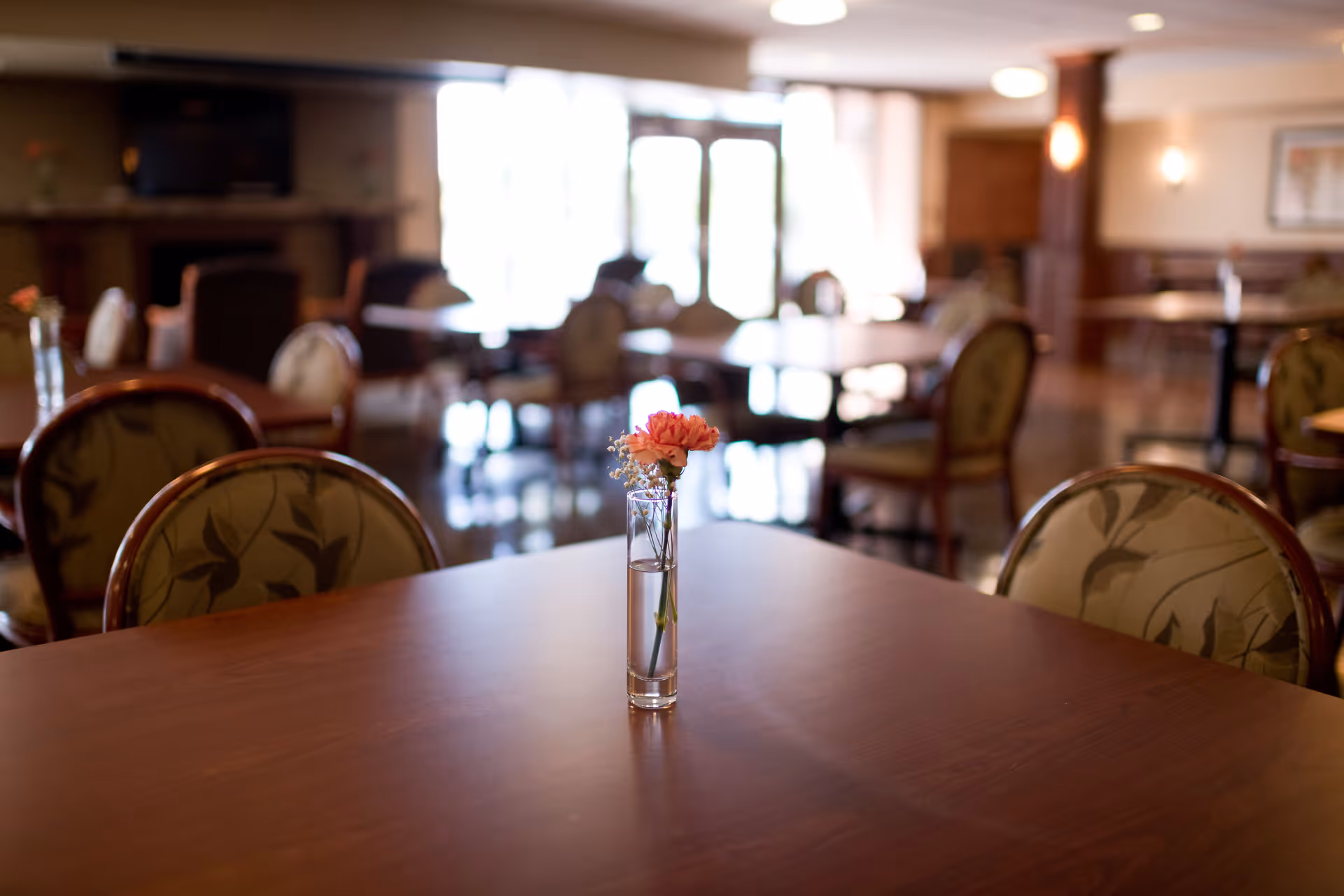 A dining area with wooden tables and cushioned chairs featuring floral patterns. A small glass vase with a pink flower and some baby's breath is placed on the table in the foreground. The background shows more tables and chairs with soft lighting and large windows letting in natural light.