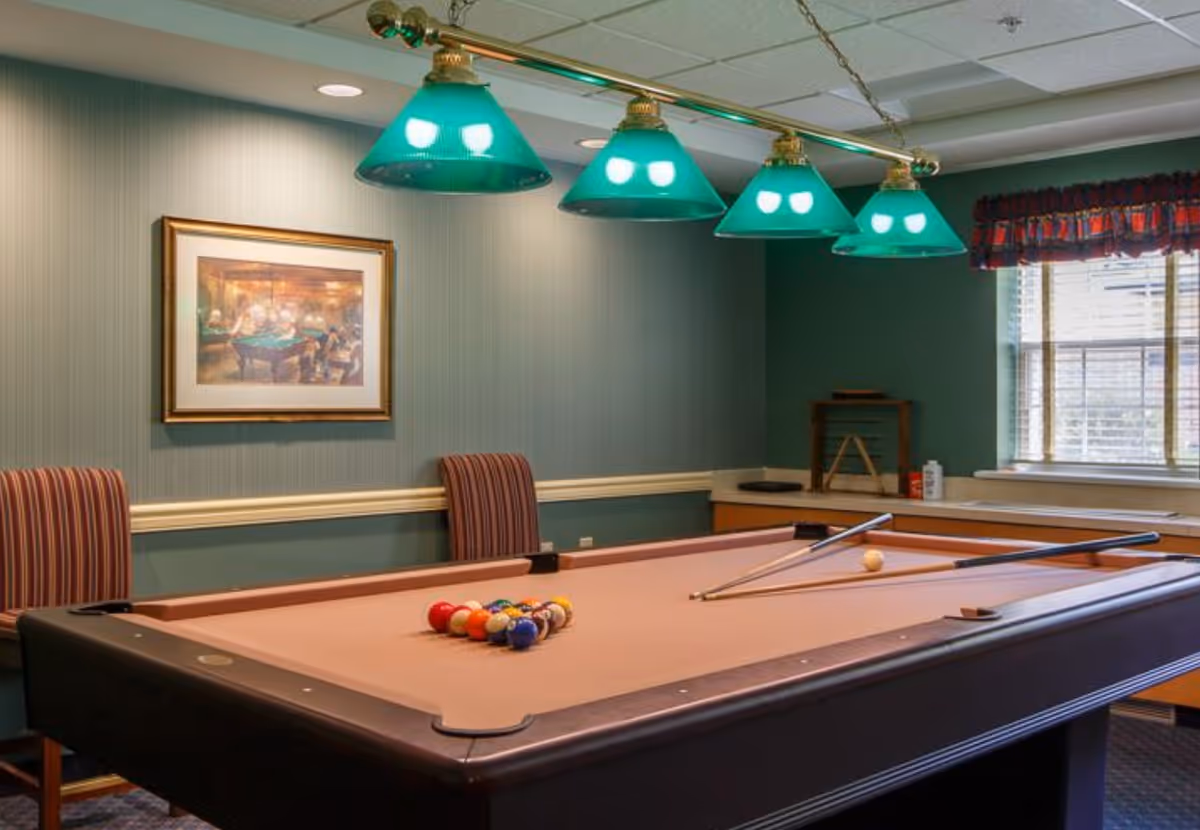 Interior room with a pool table set up for a game, two pool cues resting on the table, green hanging lights above, two striped chairs against the wall, a framed painting, and a window with blinds and a valance.
