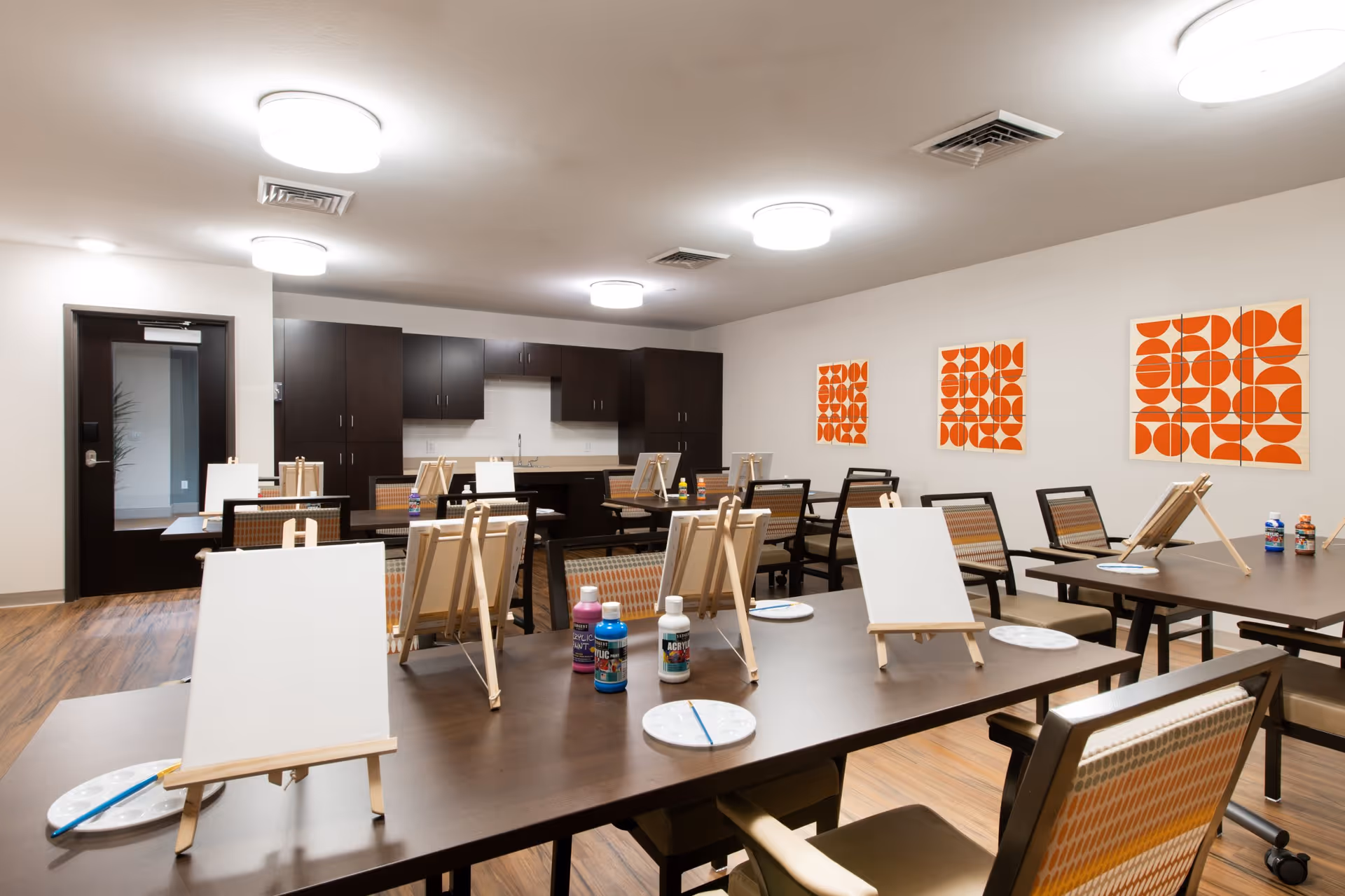A bright activity room with multiple tables and chairs arranged for painting. Each table has small easels with blank canvases, paint bottles, and palettes. The room has wooden flooring, white walls with orange geometric artwork, and dark wood cabinets along the back wall.