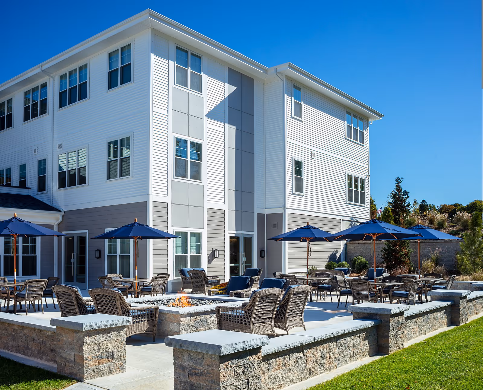 Outdoor patio area at Wingate Residences at Haverhill featuring multiple wicker chairs and tables with blue umbrellas, a fire pit in the center, and a three-story white and gray building in the background under a clear blue sky.