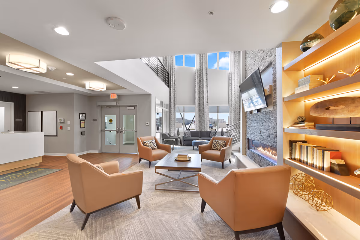 Modern senior living facility lobby with four tan armchairs around a coffee table, fireplace, TV, and built-in shelving.