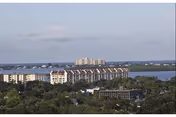 View of a waterfront area with multiple residential buildings and greenery in the foreground under a partly cloudy sky.