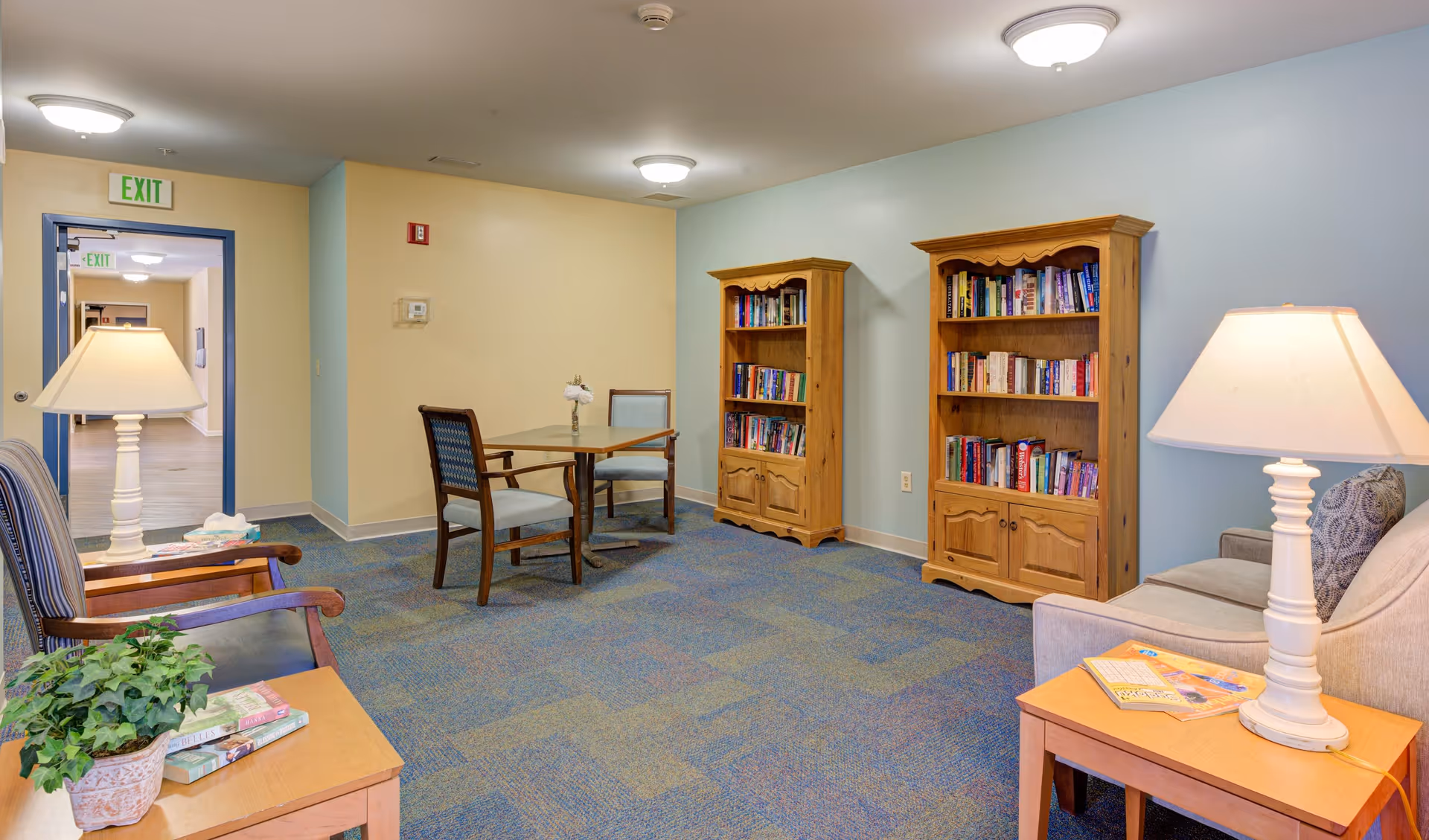 A cozy senior living community common area with two wooden bookshelves filled with books against a light blue wall. There is a small table with two chairs in the center, a beige couch with a lamp on a side table, and a wooden chair with a cushion. The floor is carpeted in blue and green patterns, and there is an exit door visible in the background.