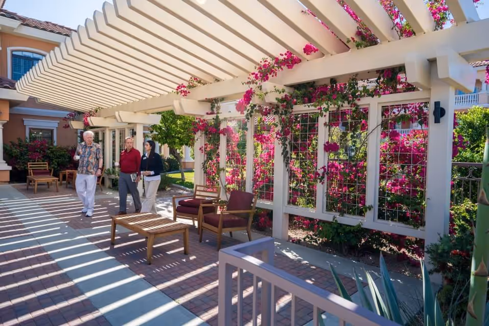 Three people walking and talking under a white pergola with pink flowering vines at a senior living facility patio area with outdoor seating and greenery.