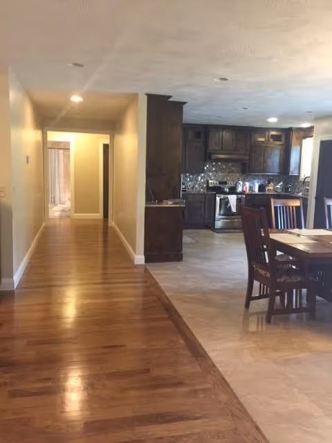 Interior view of a senior care facility showing a hallway with wooden flooring leading to a kitchen and dining area. The kitchen has dark wooden cabinets, a tiled backsplash, and stainless steel appliances. The dining area features a wooden table with chairs. The ceiling has recessed lighting.
